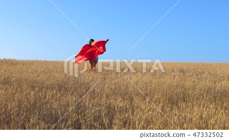 young girl in red raincoat plays in superhero runs across field of yellow wheat against a blue sky 47332502