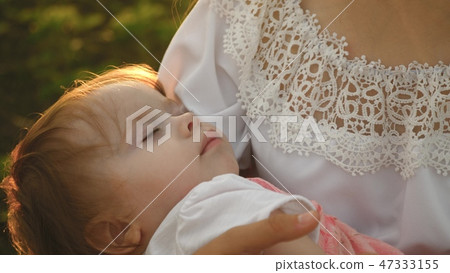 a small child falls asleep in arms of the mother, mother and daughter are walking in the summer park a small child falls asleep in arms of the mother, mother and daughter are walking in the summer park 47333155