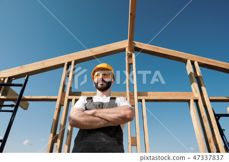 The man is a builder on the roof of a wooden frame house. The man is a builder on the roof of a wooden frame house. 47337837