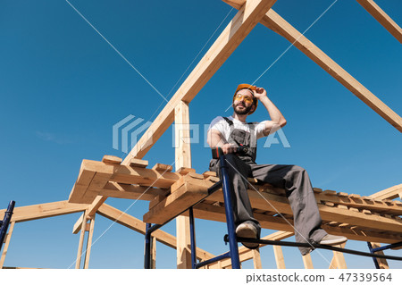 The man builder sits on the edge of the roof of the frame house, in a yellow helmet and gray 47339564