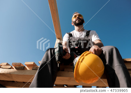 The man builder sits on the edge of the roof of the frame house, in a yellow helmet and gray 47339905