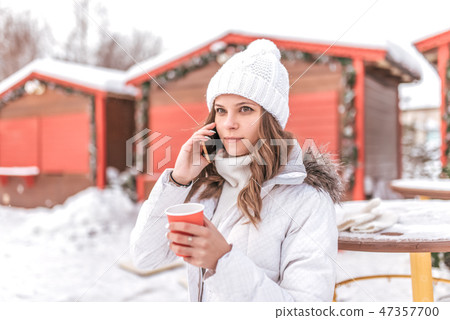 A beautiful and young girl is calling by phone, holding a cup with hot coffee or tea in her hands A beautiful and young girl is calling by phone, holding a cup with hot coffee or tea in her hands 47357700