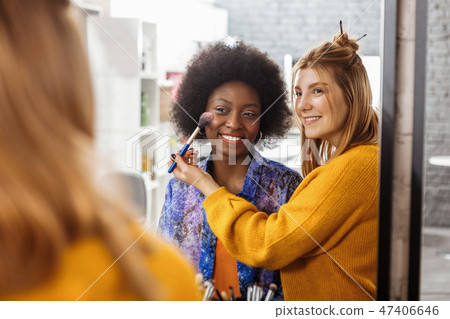 Fair-haired smiling stylist trying on a new blusher swatch on the dark-skinned model Fair-haired smiling stylist trying on a new blusher swatch on the dark-skinned model 47406646
