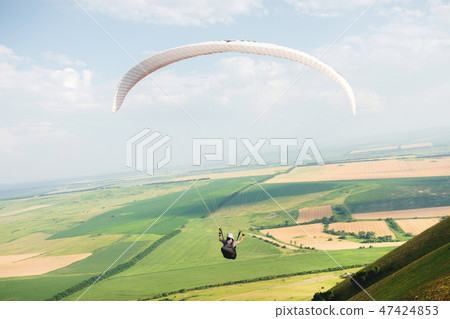 Professional paraglider in a cocoon suit flies high above the ground against the sky and fields 47424853