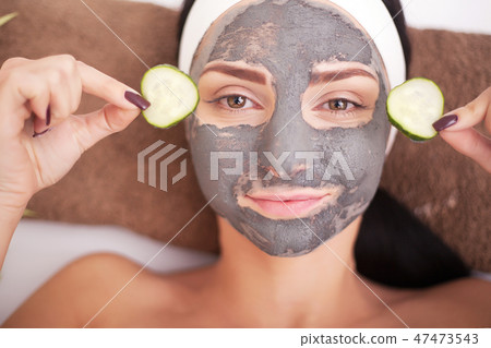 Beautiful young woman is getting facial clay mask at spa, lying with cucumbers on eyes Beautiful young woman is getting facial clay mask at spa, lying with cucumbers on eyes 47473543