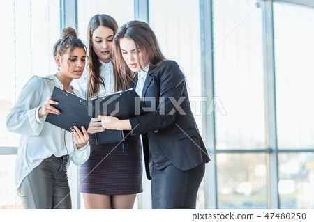 Group of friendly three multiethnic business people stand in office hall, hold papers, study Group of friendly three multiethnic business people stand in office hall, hold papers, study 47480250