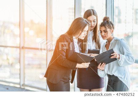 Group of friendly three multiethnic business people stand in office hall, hold papers, study Group of friendly three multiethnic business people stand in office hall, hold papers, study 47480252