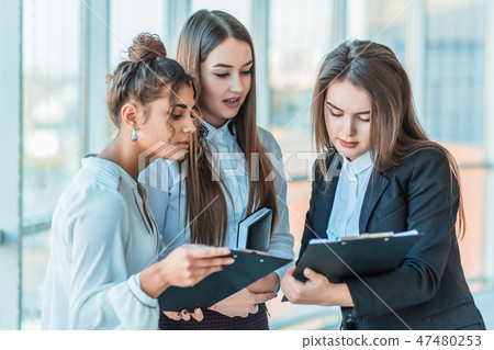 Group of friendly three multiethnic business people stand in office hall, hold papers, study Group of friendly three multiethnic business people stand in office hall, hold papers, study 47480253