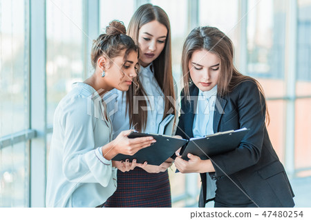 Group of friendly three multiethnic business people stand in office hall, hold papers, study Group of friendly three multiethnic business people stand in office hall, hold papers, study 47480254