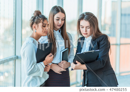 Group of friendly three multiethnic business people stand in office hall, hold papers, study Group of friendly three multiethnic business people stand in office hall, hold papers, study 47480255