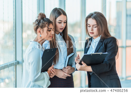 Group of friendly three multiethnic business people stand in office hall, hold papers, study Group of friendly three multiethnic business people stand in office hall, hold papers, study 47480256