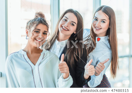 Three young business girls in the office. Dressed in classic clothing style, showing thumbs up with Three young business girls in the office. Dressed in classic clothing style, showing thumbs up with 47480315