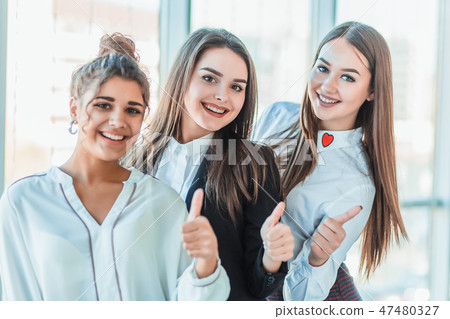 Three young business girls in the office. Dressed in classic clothing style, showing thumbs up with Three young business girls in the office. Dressed in classic clothing style, showing thumbs up with 47480327