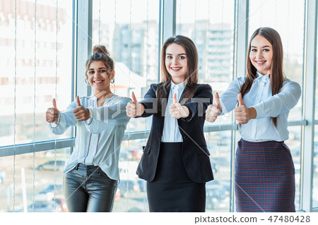 Three young business girls in the office. Dressed in classic clothing style, showing thumbs up Three young business girls in the office. Dressed in classic clothing style, showing thumbs up 47480428