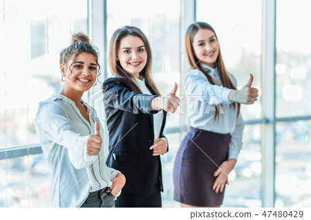 Three young business girls in the office. Dressed in classic clothing style, showing thumbs up Three young business girls in the office. Dressed in classic clothing style, showing thumbs up 47480429