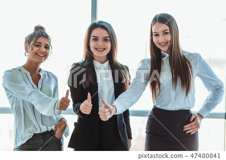 Three young business girls in the office. Dressed in classic clothing style, showing thumbs up Three young business girls in the office. Dressed in classic clothing style, showing thumbs up 47480841