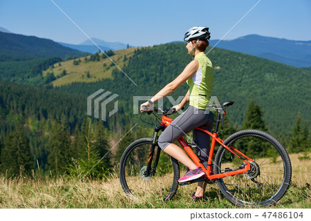 Female biker in helmet standing with red bicycle on grassy hill 47486104