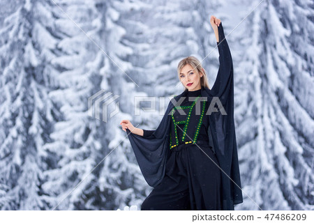Attractive woman in dark long fantasy outfit outdoors in deep snow on sunny winter day. 47486209