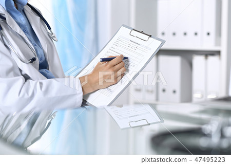 Female doctor filling up medical form on clipboard, closeup. Reflecting glass table is a physician 47495223