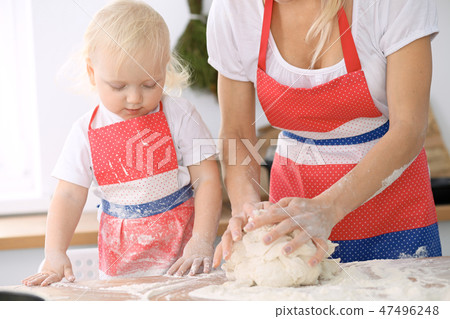 Little girl and her blonde mom in red aprons playing and laughing while kneading the dough in 47496248