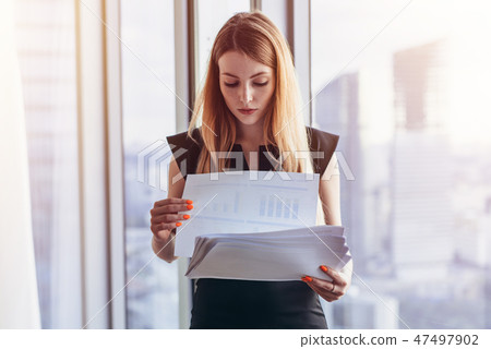Confident female director holding documents standing in modern office against panoramic window Confident female director holding documents standing in modern office against panoramic window 47497902
