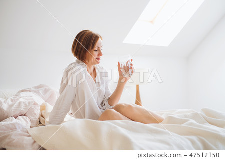 A young woman with night shirt sitting indoors on bed in the morning, holding water. A young woman with night shirt sitting indoors on bed in the morning, holding water. 47512150