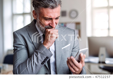 A frustrated businessman with smartphone standing in an office, reading bad news. 47512237