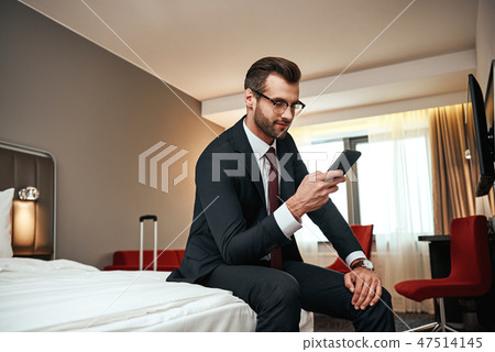 Businessman in business suit sitting with smartphone on bed at hotel room 47514145
