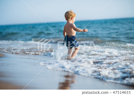 A small toddler boy with shorts walking in water on beach on summer holiday. A small toddler boy with shorts walking in water on beach on summer holiday. 47514655