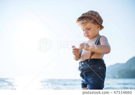 A small toddler boy with hat and shorts standing on beach on summer holiday. 47514660