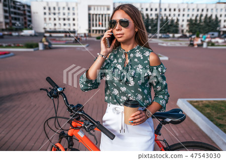 Beautiful girl brunette woman standing in the park behind the back of the bike. Phone in hand a mug Beautiful girl brunette woman standing in the park behind the back of the bike. Phone in hand a mug 47543030