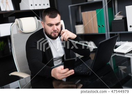 Busy young man talking on phone and looking at laptop in office Busy young man talking on phone and looking at laptop in office 47544979