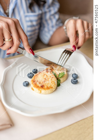 Beautiful young woman cuts pancakes with a knife. Light breakfast with coffee near window in a cafe. Beautiful young woman cuts pancakes with a knife. Light breakfast with coffee near window in a cafe. 47560525