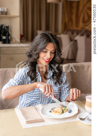 Beautiful young woman cuts pancakes with a knife, enjoying coffee cappuccino or latte in a glass cup 47560530