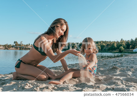 Beautiful young woman mom playing on shore of a lake with sand, next to a small child is a daughter Beautiful young woman mom playing on shore of a lake with sand, next to a small child is a daughter 47566034