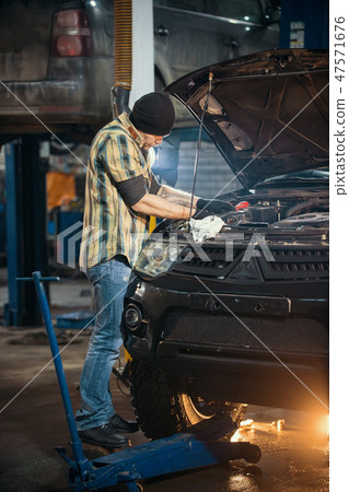 Car service. Brutal mechanic man standing by the car with open hood and inspects the engine Car service. Brutal mechanic man standing by the car with open hood and inspects the engine 47571676