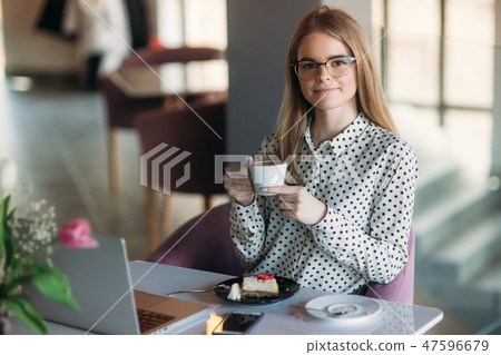 Girl eating cake in a cafe. Blonde girl in polka-dot shirt with laptop. Drinking cappuccino Girl eating cake in a cafe. Blonde girl in polka-dot shirt with laptop. Drinking cappuccino 47596679