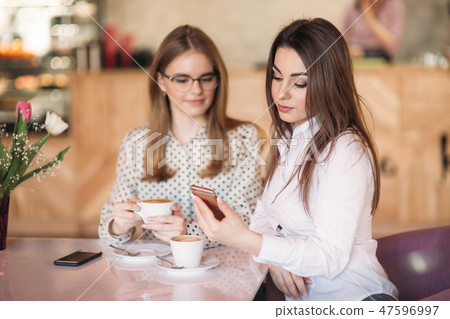 Two girls sitting in a cafe drinking cappuccino and using smartphone. Modern girl Two girls sitting in a cafe drinking cappuccino and using smartphone. Modern girl 47596997