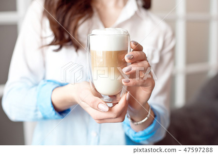 Close-up of female hands in office clothes holding a glass cup of coffee with soy milk during a Close-up of female hands in office clothes holding a glass cup of coffee with soy milk during a 47597288