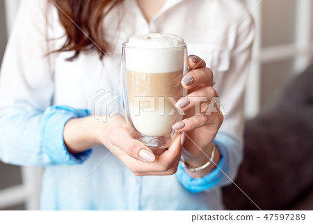 Close-up of female hands in office clothes holding a glass cup of coffee with soy milk during a Close-up of female hands in office clothes holding a glass cup of coffee with soy milk during a 47597289