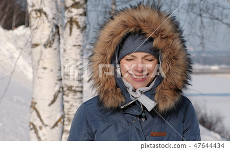 Close-up woman in blue down jacket with fur hood frown from the sun and smiles,in winter Park. 47644434