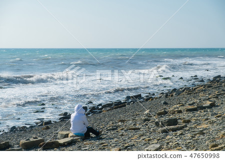 Unrecognizable woman in white jacket sitting on the seafront in stone. Winter at sea. 47650998