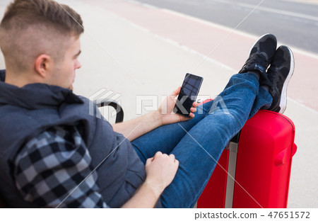 Travel, tourism and people concept - handsome man sits and put his feet on suitcase, watching 47651572