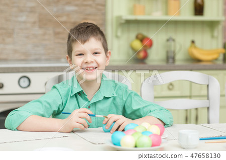 Little blond kid boy coloring eggs for Easter holiday in domestic kitchen 47658130