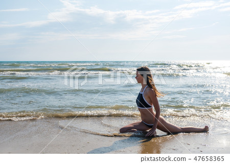 Woman bruenette with long hairs in blue bikini is stretching on the seashore. Side view. 47658365