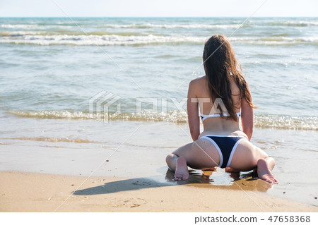 Unrecognizable woman bruenette with long hairs in a bikini sits on the sand beach. Back view. 47658368