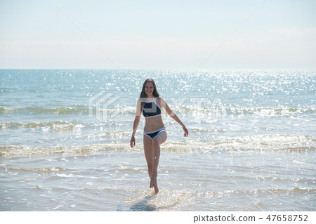 Brunette woman with long hair in blue bikini walks along the sea on a sunny day and smiles. 47658752