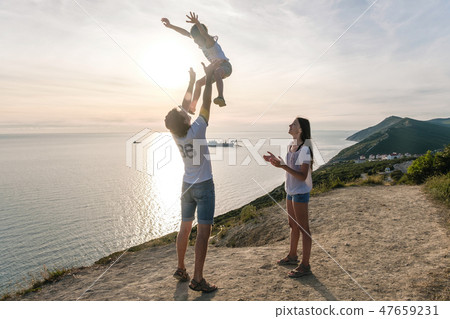 Dad throws his son in the air, and mom looks at them on the mountain with seascape. Family trip. Dad throws his son in the air, and mom looks at them on the mountain with seascape. Family trip. 47659231