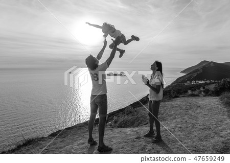 Dad throws his son in the air, and mom looks at them on the mountain with seascape. BW 47659249