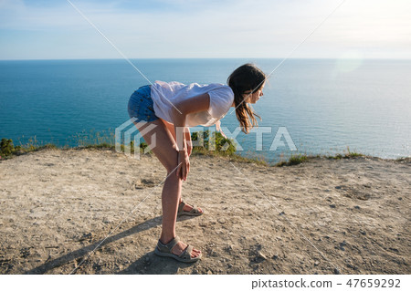 Woman brunette in light blouse and denim shorts stretching on the hill with seascape. Side view. 47659292
